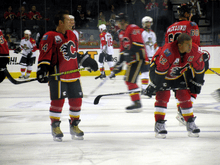 A player in full uniform but without a helmet stands to the right of a teammate who is crouched over. Both players are looking to their left as several others skate in the background.
