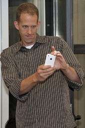 The portrait of a man holding a white telephone. He is wearing a striped brown shirt.