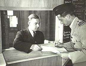 Two men in military uniforms, one wearing a forage cap and filling out a form