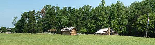A panoramic view of the historic site showing two cabins and a well, as well as a flagpole bearing the British flag