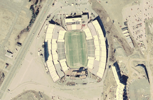 An aerial view of a large sports stadium with a four-lane road next to it on the left and the beginnings of a similar structure at lower right