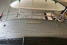 Photograph showing part of a ship's curved hull, bearing the name FRAM. A small lifeboat has been slung above the ship's rail.