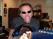 Musician Frank Klepacki holding the neck of his guitar to the camera wearing sunglasses, collectibles on shelves in the background