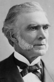 A black and white, head and shoulders photographic portrait of an elderly caucasian gentleman with greying hair and white mutton chop sideburns. He is smartly dressed in a dark suit jacket and white shirt with a Gladstone collar and Kentucky style bowtie.