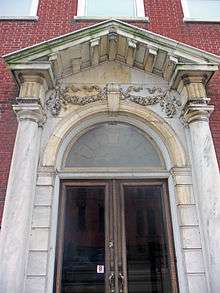An arched stone entrance on a brick background, with smooth columns on either side and metal-framed glass double doors topped by a round-arched transom with keystone and a stone pediment
