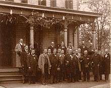 A formative shot taken in front of an ornate colonial-style home. A middle-aged gentleman with thinning hair stands at center of a group of about fifteen men.