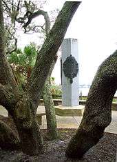A cement monument, a pentagon-shaped column with bronze plaques adorned with the fleur-de-lis, surrounded by oak trees in the foreground, palmettos to the left all overlooking the river