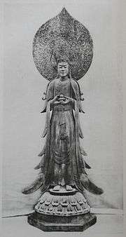 Front view of a standing statue with flowing robes holding a small container in front of her body. There is a large halo behind the head of the statue.