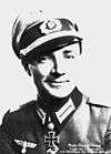 Black-and-white portrait of a smiling man wearing a peaked cap, military uniform with an Iron Cross displayed at his neck.