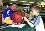 An older man, wearing a purple and yellow jacket is holding a basketball, handed out by a kid who is standing in front of him.