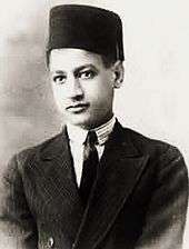 A boy wearing a jacket, a white shirt with a black tie and a fez on his head