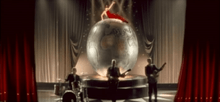 The stage concert house, with red curtains on the side and grey ones in the background. Atop a scenographic Earth globe lies a woman in a red dress, while below it three men in suits play drums, bass and guitar.