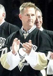 Upper body of a man with short, brown hair applauds. He is wearing a black hockey sweater with white and yellow trim with a stylized penguin logo.