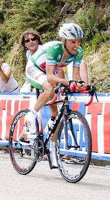 A cyclist in a green and white jersey with red trim, with a pained look on his face as he strains to climb a hill on his own. Spectators watch him from behind a barricade.