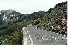 A curving, ascending road, up against a rocky hillside. There is writing in Italian on the road, a sign on the roadside, and further mountaintops visible in the background.