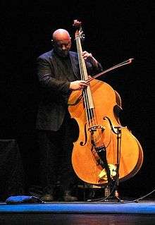 A bald white man stands playing the double bass in front of a low microphone.