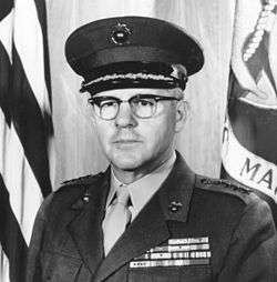 A stocky Caucasian man with brown hair and thick glasses in military uniform in front of a flag