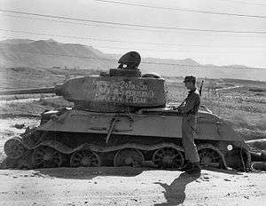 Soldiers stand around a destroyed tank with writing on it reading "Knocked out 20 July 1950 with the supervision of Maj. Gen. W.F. Dean"