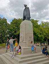 A dark brown statue of a man in 18th-century military uniform, including tricornered hat, on a light tan stone pedestal. Around its base people dressed primarily in T-shirts and shorts are lounging. There are trees a short distance beyond