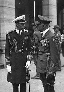Photograph of two men in formal military uniform. The man on the left is a naval officer's uniform, and has a cap, white gloves, a ceremonial sword and has medal ribbons on his chest. The man on the right is also in a military officer's uniform, and is wearing a cap, leather gloves and medals. He is carrying a ceremonial sword in his right hand. Other men in military uniform can be seen in the background.