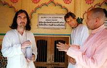 Three men in their early fifties, one on the left, wearing a white robe and holding a bottle of water with both hands, and two on the right, one in a white robe and one in a pink robe. On the wall behind them is something written in Sanskrit, both in Roman characters and in Sanskrit characters.