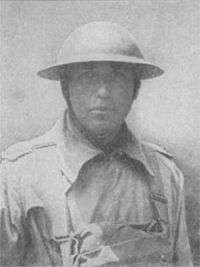 Head and shoulders of a man in a military coat, buttoned up to the neck, with a light pack strapped to his chest. He is wearing a metal helmet with a wide brim, its chin strap tightly cinched.