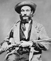 Black and white image of a bearded man with a hat, wearing a jacket over a suit and tie; he is seated and holding a rifle across his lap.
