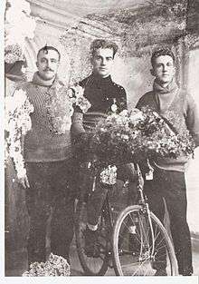 Three men standing next to each other. The man in the middle is sitting on a bicycle with many flowers. The picture is slightly damaged, as if it is an old photograph.