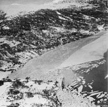 Two ships close to the shore of a body of water near steep snow-covered hills. Much of the body of water is covered by sheets of ice.