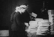 A black-and-white film still in the four corners. Three men in the center stand by a table on the right stacked with thousands of sheets of paper.