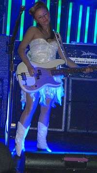 Ginger Reyes—a Caucasian woman in her late 20s with long brunette hair—plays bass guitar on stage wearing a white dress and white cowboy boots.