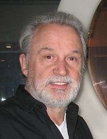 Headshot of a bearded man wearing an unbuttoned black collared shirt over a white T-shirt.