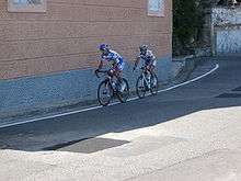 Two cyclists, both wearing white and blue jerseys, one with yellow trim, ride on an empty road beside a red and blue building.