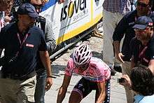 A road racing cyclist, in a pink jersey with a pink helmet, riding among men wearing blue polo shirts and pink lanyards around their necks. Partly visible at the bottom of the frame is a woman holding a camera.