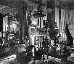 A black and white photograph of a well-decorated room with a globed hanging light fixture. On the left is an entryway to a neighboring room, similarly decorated