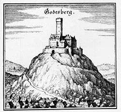 A medieval fortress stands on the peak of a hill, dominating the valley below it, and the river passing it in the distance; the fortress has a central keep (tower), and an elaborate ring of crenelated walls and narrow windows.  A road twists around the mountain to the gate.