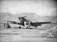Three men stand near a propeller aircraft on a runway. There are tall mountains in the background.