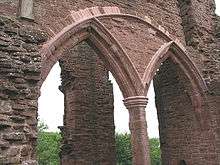 Part of a castle, with a huge semi-circular arch containing two smaller Norman arches dominating the picture. Through the arches, a ruined pillar can just be made out.