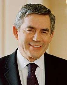 Head and shoulders of a smiling man in a dark suit and spotted tie with dark, greying hair and rounded face with square jaw