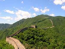A high defensive wall with watch towers running through a mountain landscape.