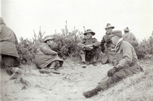 Four caucasian soldiers in uniform sitting on the ground talking. Three of the men are wearing slouch hats, while the fourth is wearing a cap.