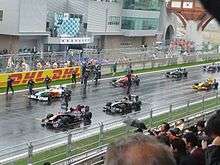 Seven Formula One cars are shown stationary on a wet racing track.