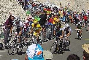 Four cyclists riding up a mountain, chased by two more cyclists. On each side of the road, spectators cheer on the riders.