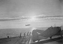 Black and white photograph of a single-engined monoplane on the wooden deck of an aircraft carrier with four men walking along the deck towards the aircraft. Another aircraft is flying above the sea in the background to the photo.