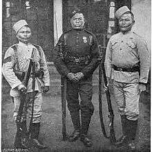 Three men in 1800s-style military uniforms stand holding rifles.