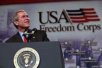 President George W. Bush stands at a podium with a large banner behind him that says "USA Freedom Corps".