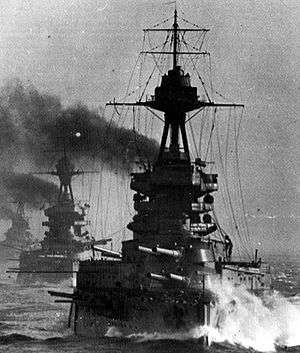 A large gray battleship steams in choppy seas; thick black smoke billows from its funnels. Two battleships are directly behind