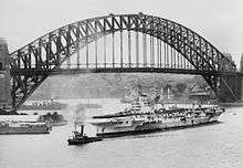 Black and white photo of a World War II-era aircraft carrier in front of a steel through arched bridge. Several other ships are visible near the aircraft carrier
