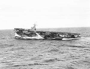 Black and white photograph of an aircraft carrier at sea. The stern of the ship is much lower in the water than the bow.