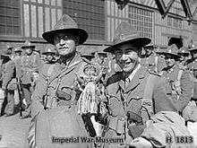 Two Maori men wearing military uniforms smile at the camera, surrounded by other soldiers in front of a building
